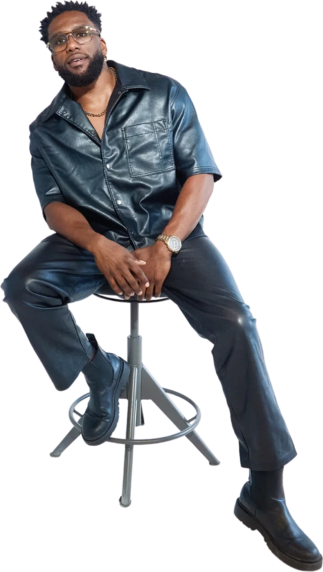 Chamalo seated on a stool in a black leather shirt and trousers