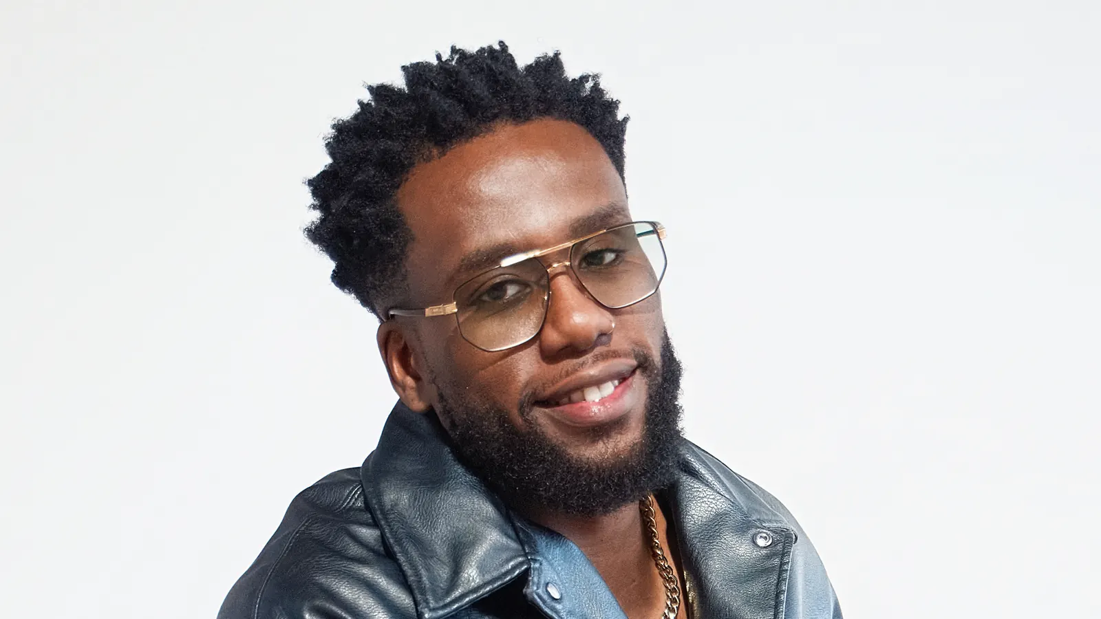 Chamalo smiling, wearing glasses and a black leather jacket, in the studio